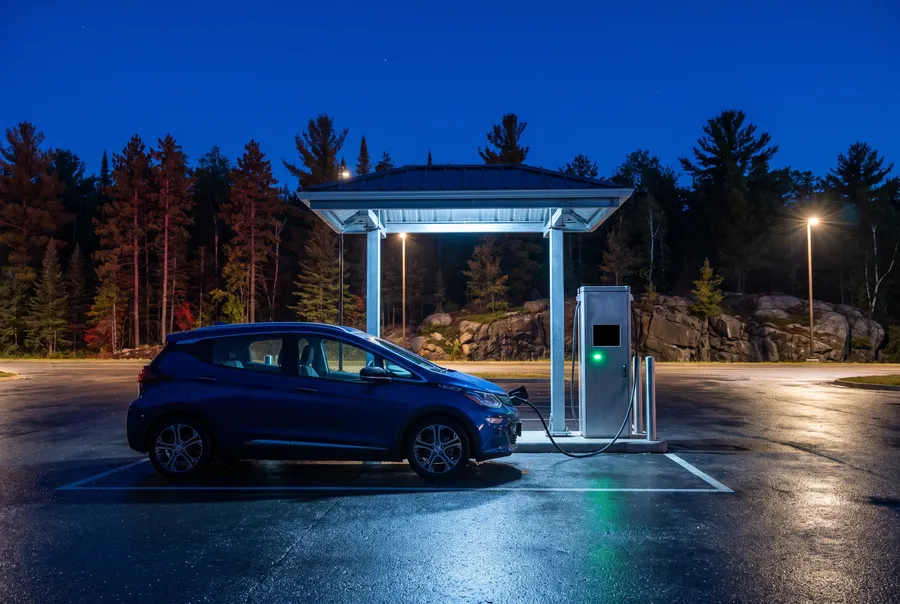 Generated photograph: a blue electric hatchback plugged into a charging post under a small canopy at night, pines and granite behind.